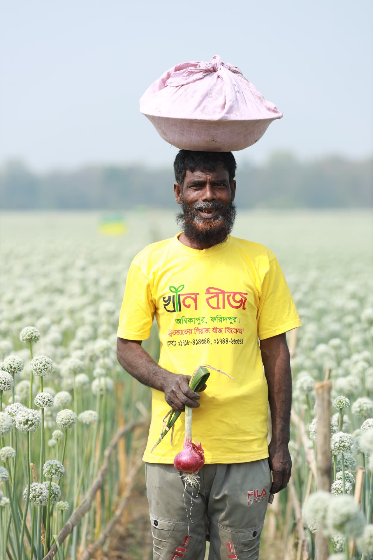 Farmer in a Khan Seed t-shirt carrying a harvest bundle and holding a red onion